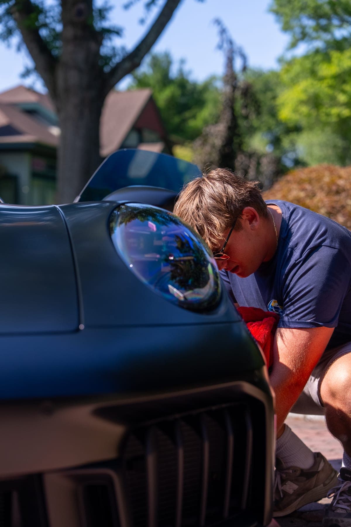 Pro Performance owner-operator bent close over the front headlight of a matte-black Porsche 911, natural sunlight, tree canopy behind.