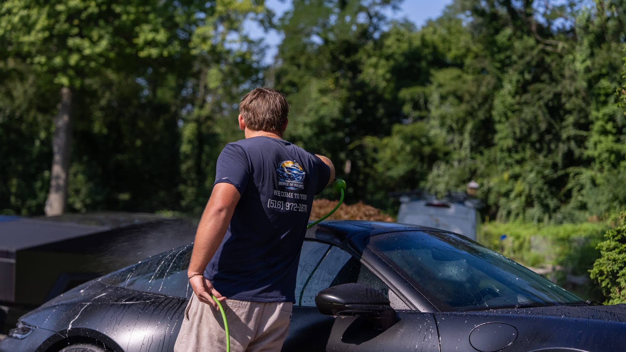 Pro Performance owner-operator in a branded shirt reading WE COME TO YOU · (516) 972-1674, hose in hand at the side of a matte-black Porsche 911 in a residential driveway.