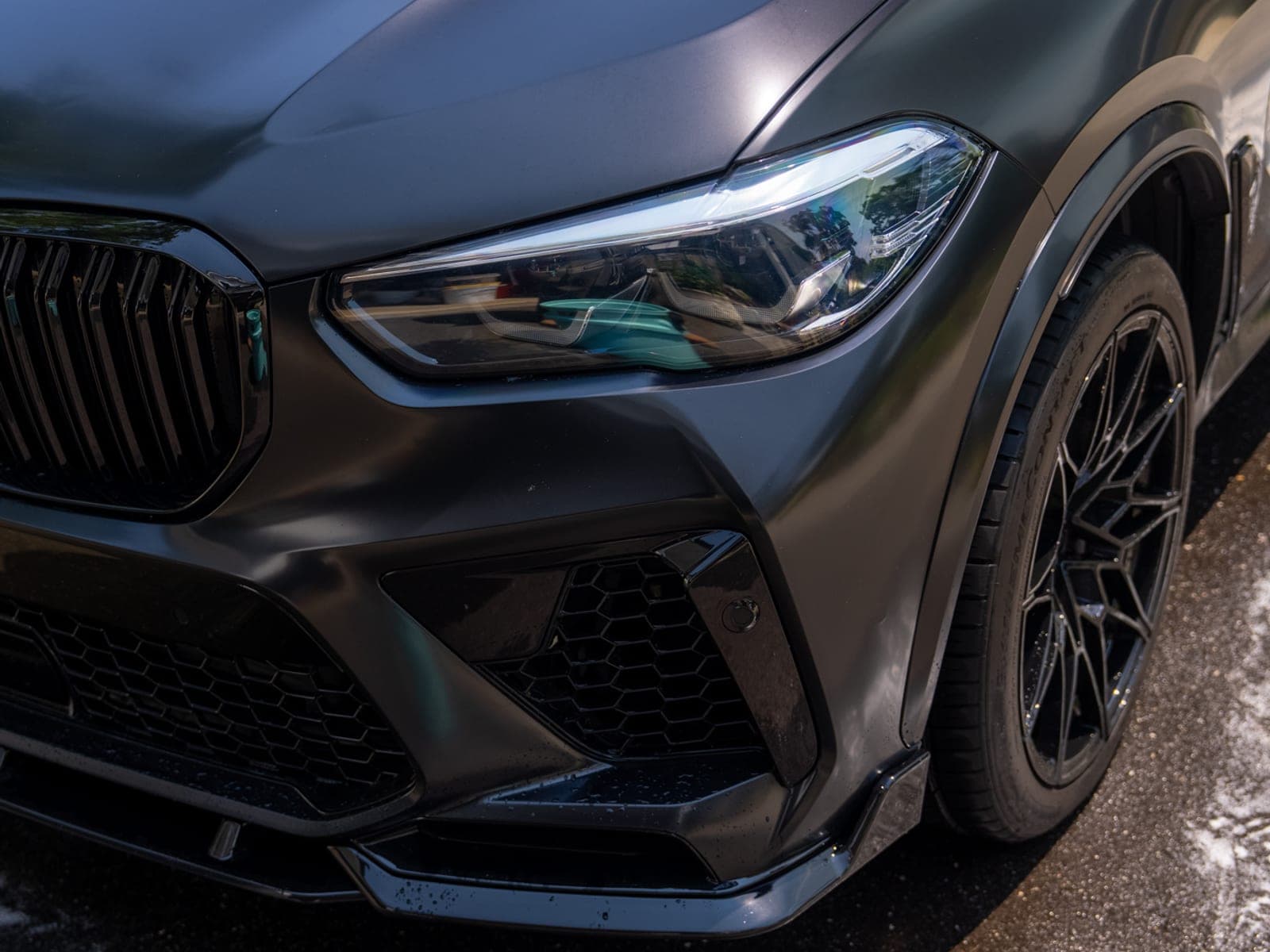 Front three-quarter view of a matte-black BMW X5M in a residential driveway, deep gloss across the fender and headlight after ceramic protection.