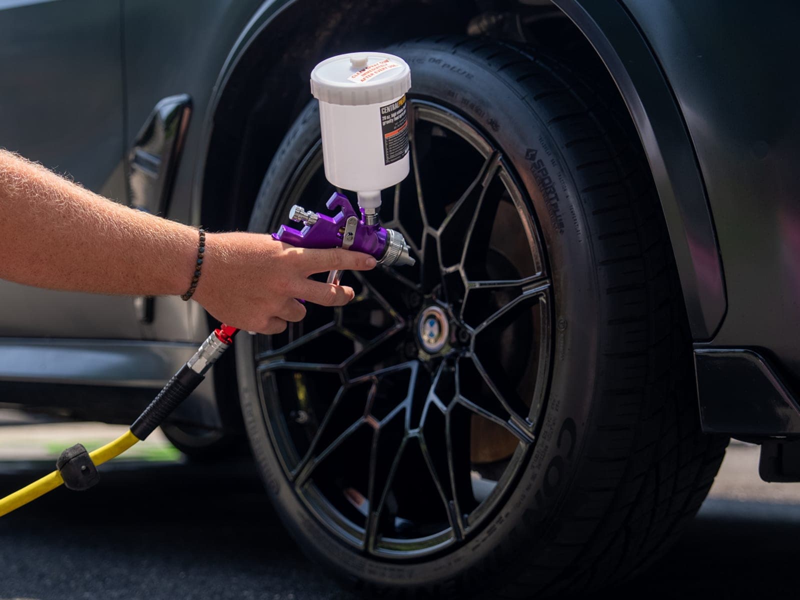 Pro Performance owner-operator's hand aiming a wheel-cleaning spray gun at a black BMW X5M wheel, yellow air hose looped on the driveway.