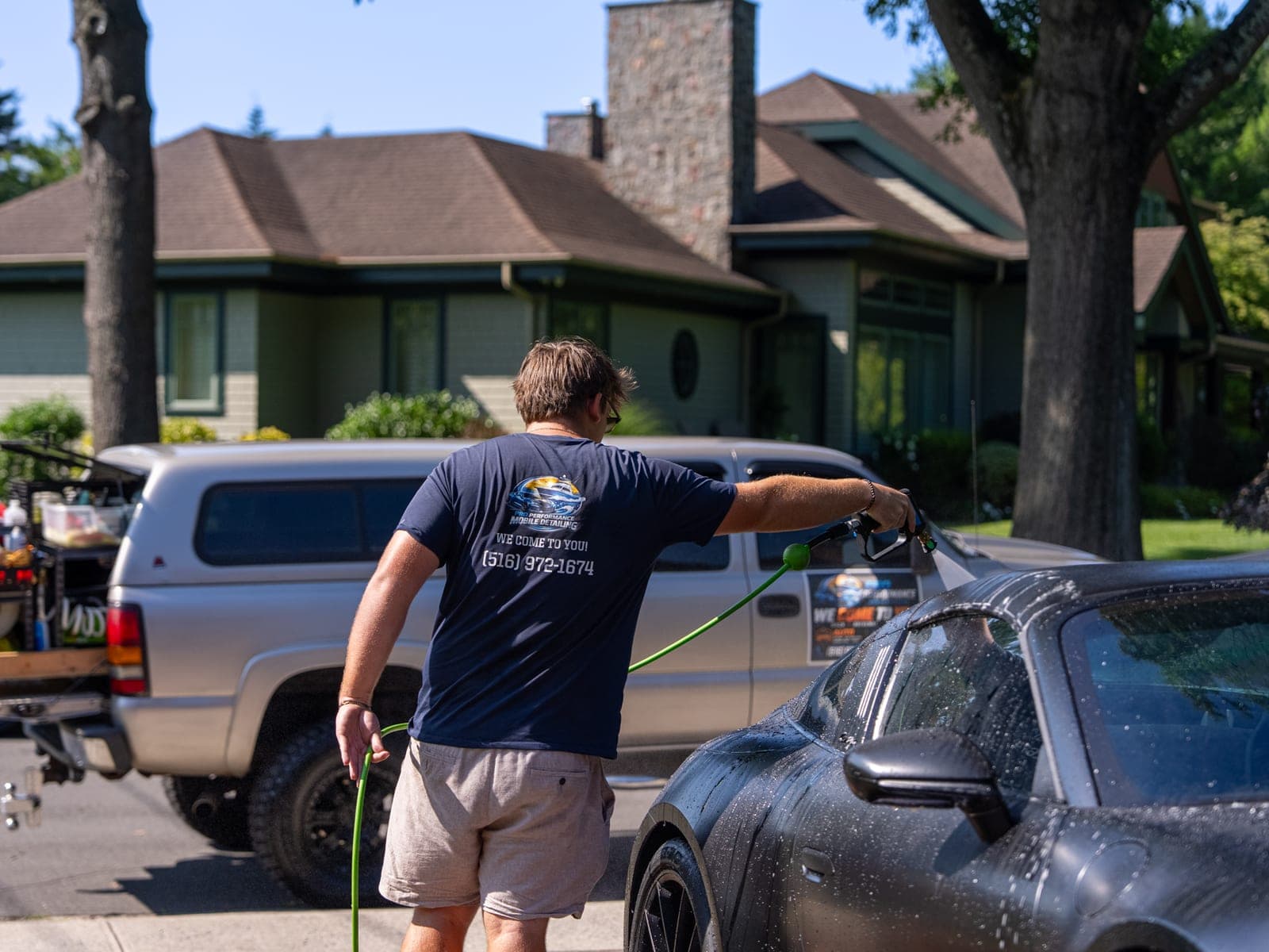 Pro Performance owner-operator in a branded 'WE COME TO YOU · (516) 972-1674' shirt holding a hose at a matte-black Porsche 911, service truck parked beside the driveway of a suburban home.