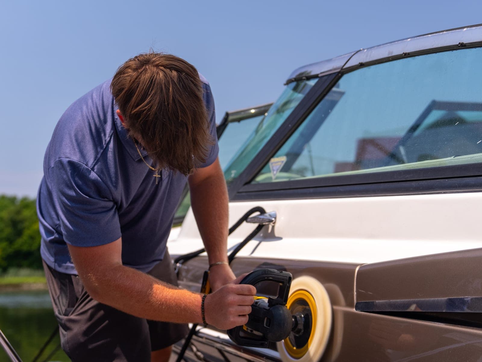 Pro Performance owner-operator running a rotary polisher across the dark hull of a cruiser at a marina slip, cream cabin windshield and water visible behind.